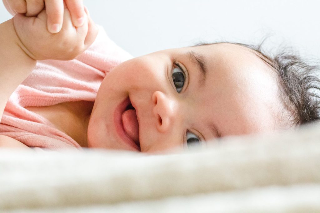 A smiling baby looking at the camera, laying on the bed