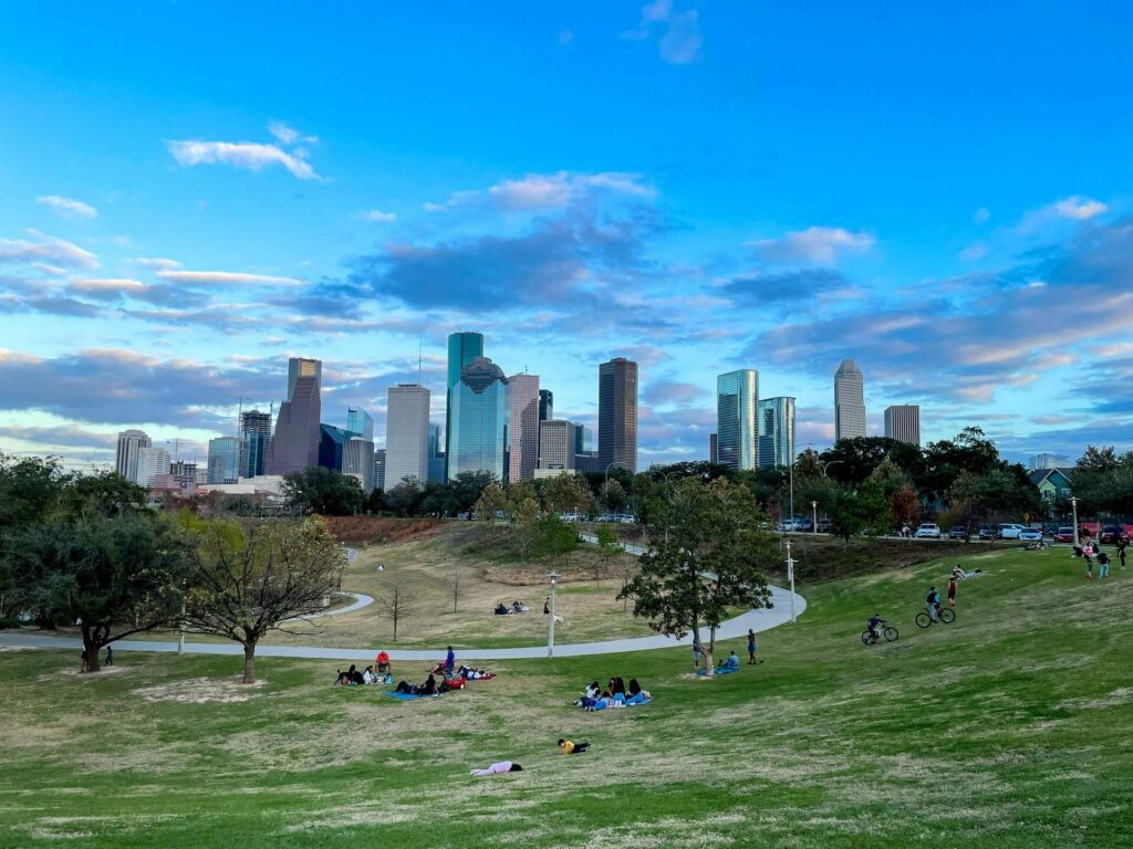 People relaxing and sitting on a grassy green park field with tall city skyscrapers visible in the background during daytime.